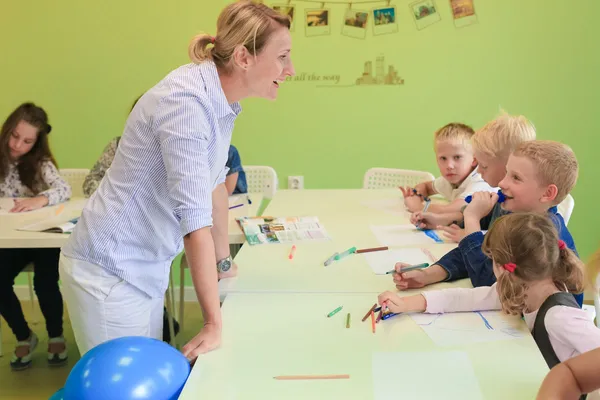 Nauczycielka pochyla się nad stołem i pomaga dzieciom podczas ćwiczenia