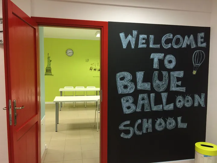 Otwarte czerwone drzwi prowadzące do sali lekcyjnej obok tablicy Welcome to Blue Balloon School