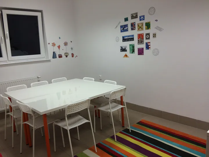 Sala lekcyjna z dużym białym stołem, krzesłami i kolorowym dywanem