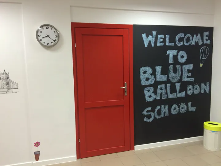 Hol szkoły z czerwoną framugą drzwi i napisem Welcome to Blue Balloon School na czarnej tablicy