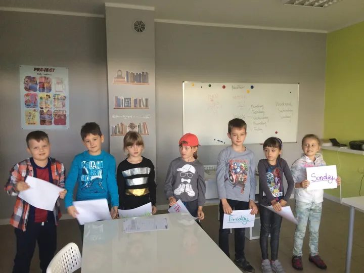 Dzieci stoją w rzędzie w sali lekcyjnej i trzymają prace w rękach